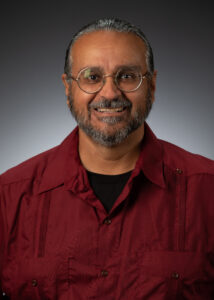 Ben V. Olguín, a man in a red guayabera shirt and glasses, smiles at the camera in front of a grey background.